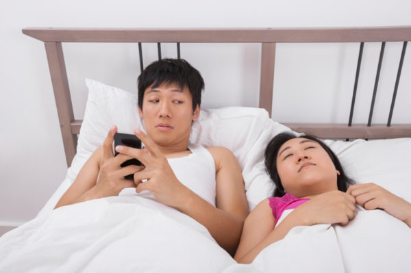 Man using cell phone while looking at woman sleeping in bed