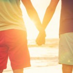 Happy gay couple holding hands watching sunset on the beach